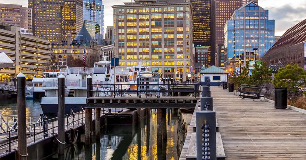 Evening waterfront view of Boston’s North End skyline, lit buildings and Old North Church spire, along a wooden boardwalk by the harbor with calm water below.